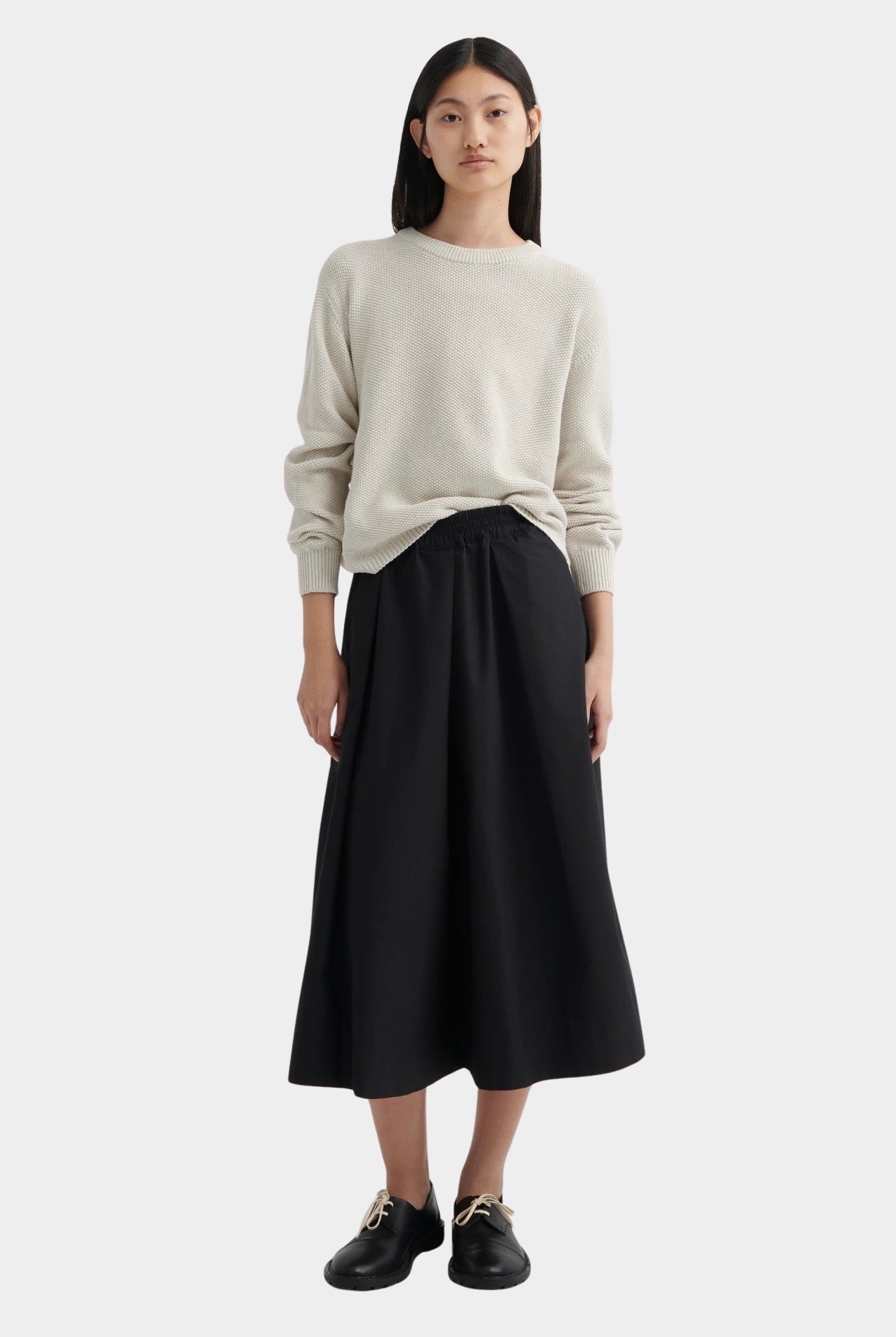 Full-length view of model wearing cream knit sweater styled with a black midi skirt and black lace-up shoes.