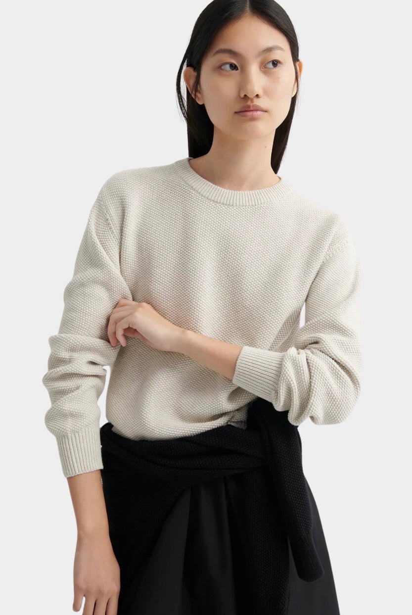 Model standing in a cream textured knit sweater with ribbed cuffs, styled with a black midi skirt.