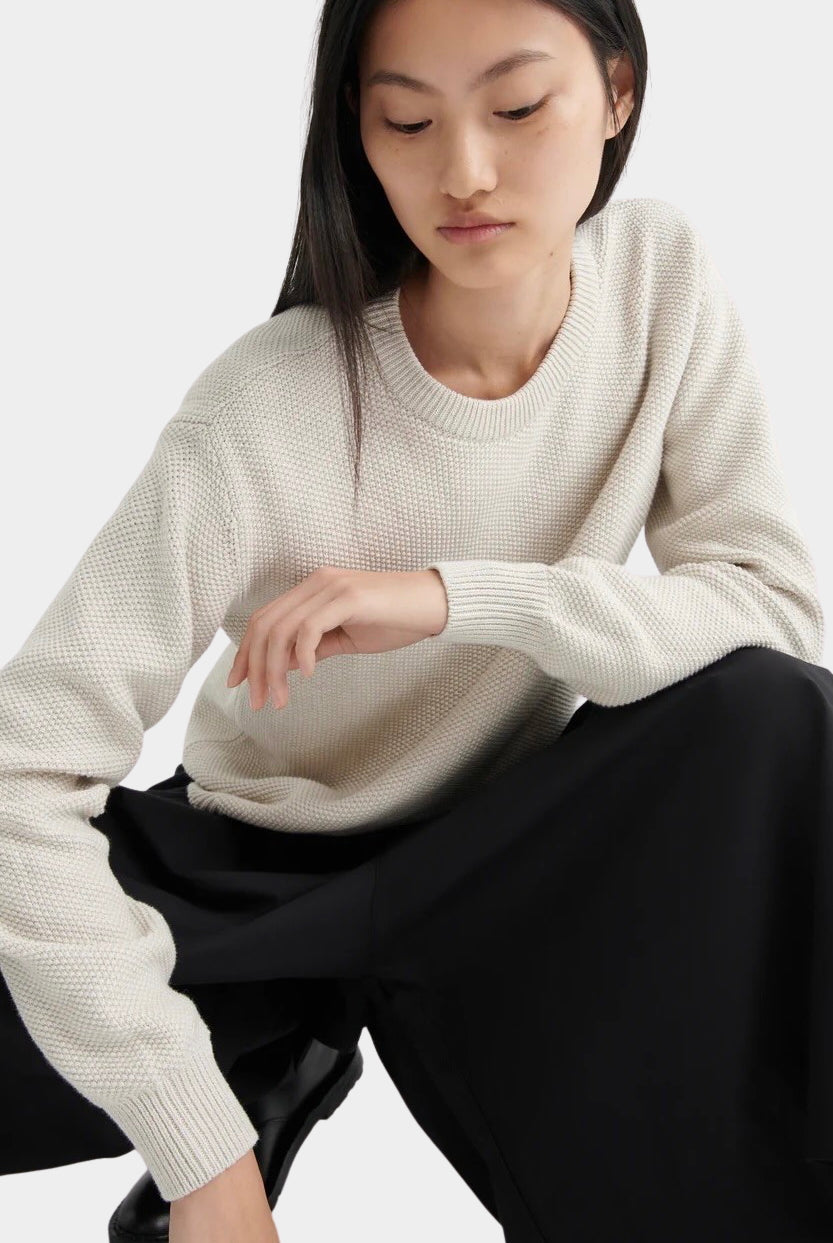Model seated wearing a textured cream crewneck sweater with black midi skirt and black leather shoes.