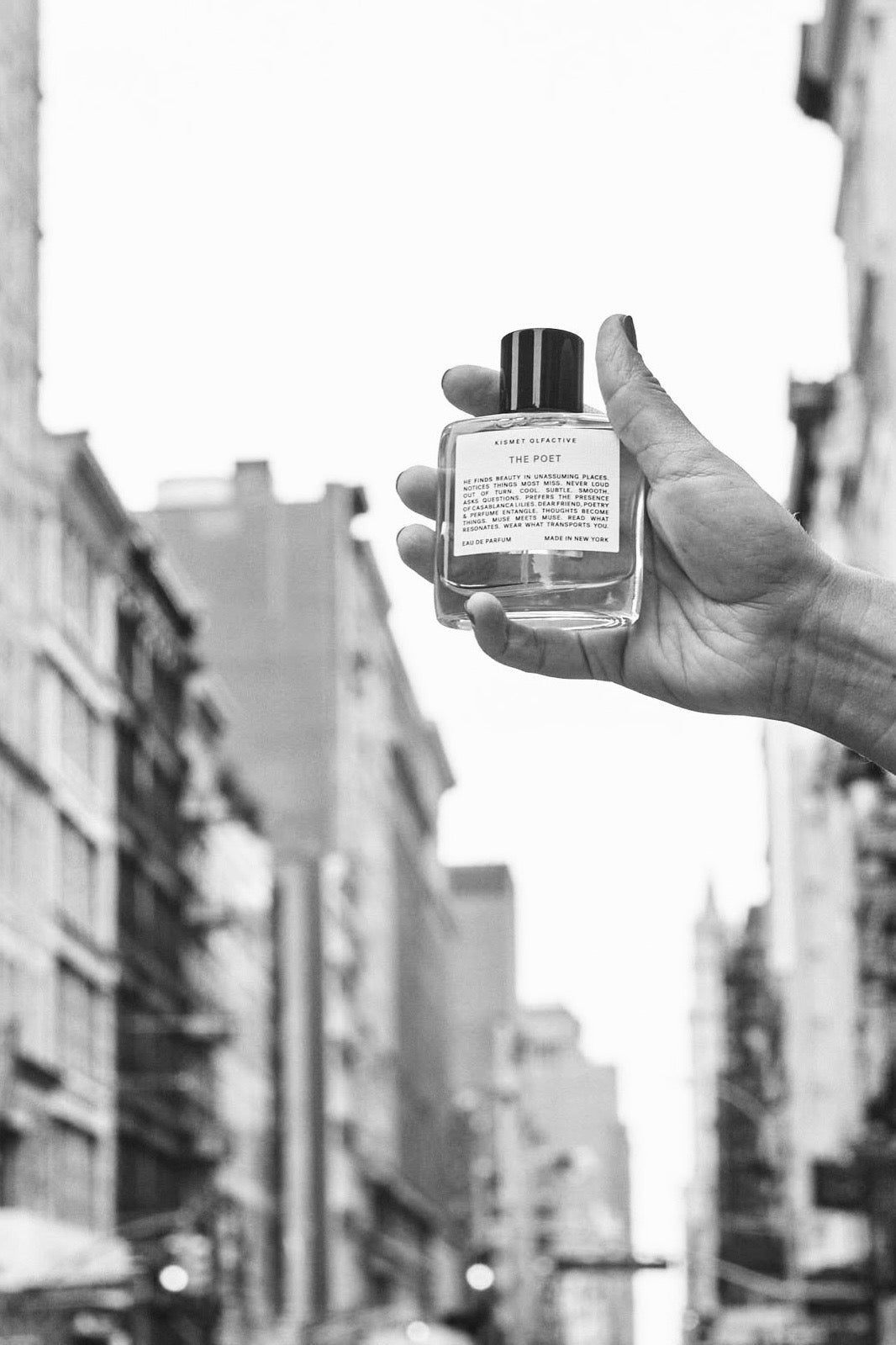 Black and white photo of hand holding Kismet Olfactive perfume bottle against city street backdrop.