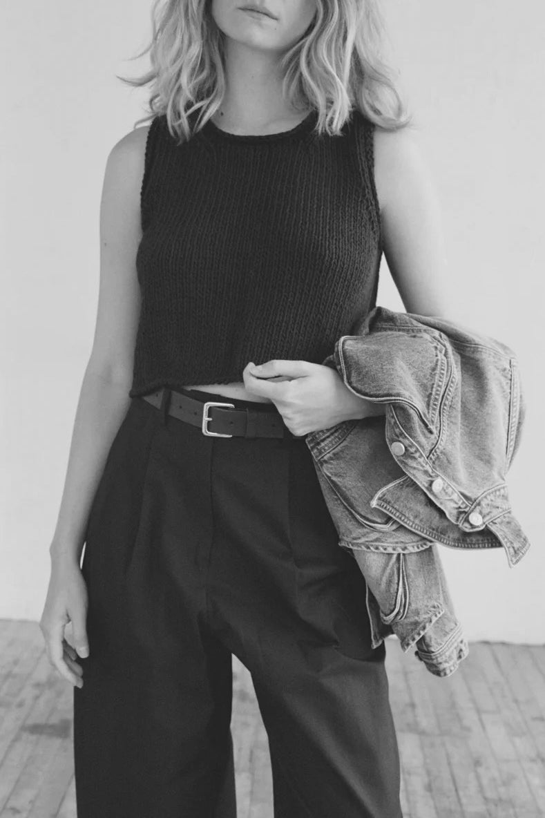 Shaina Mote clothing model wearing textured sleeveless knit top, wide-leg black trousers, and leather belt, holding denim jacket, minimalist modern womenswear style