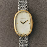 Close up of BREDA Jane Tethered mother of pearl dial, gold oval case and silver woven bracelet on gray fabric