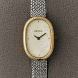 Close up of BREDA Jane Tethered mother of pearl dial, gold oval case and silver woven bracelet on gray fabric