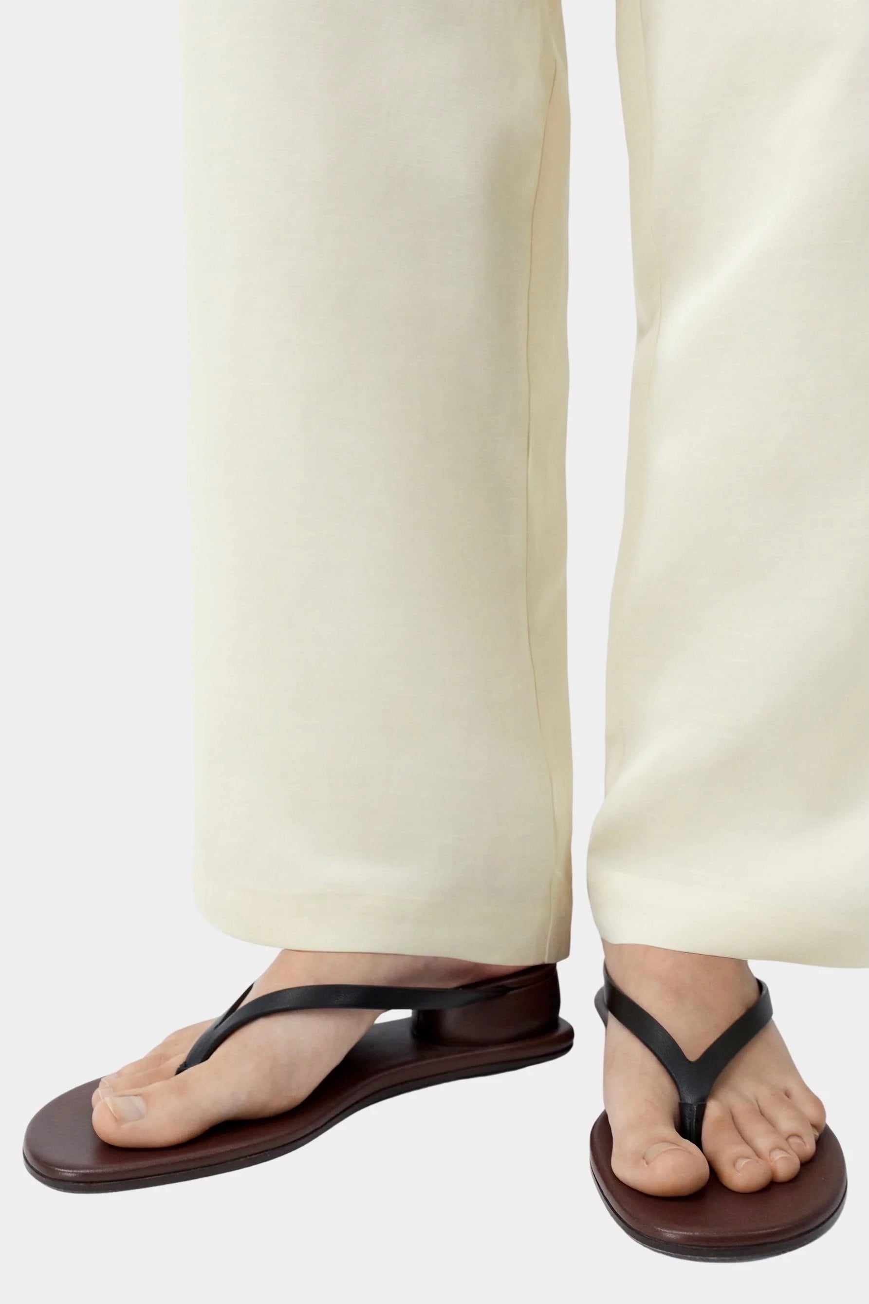 Black leather thong sandal worn with cream wide leg pants, showing low profile sole and minimal strap