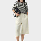 Cordera organic cotton V-neck polo in black and cream horizontal stripe with relaxed fit, short sleeves, and contrast brown collar, styled with cream culotte pants and black sandals