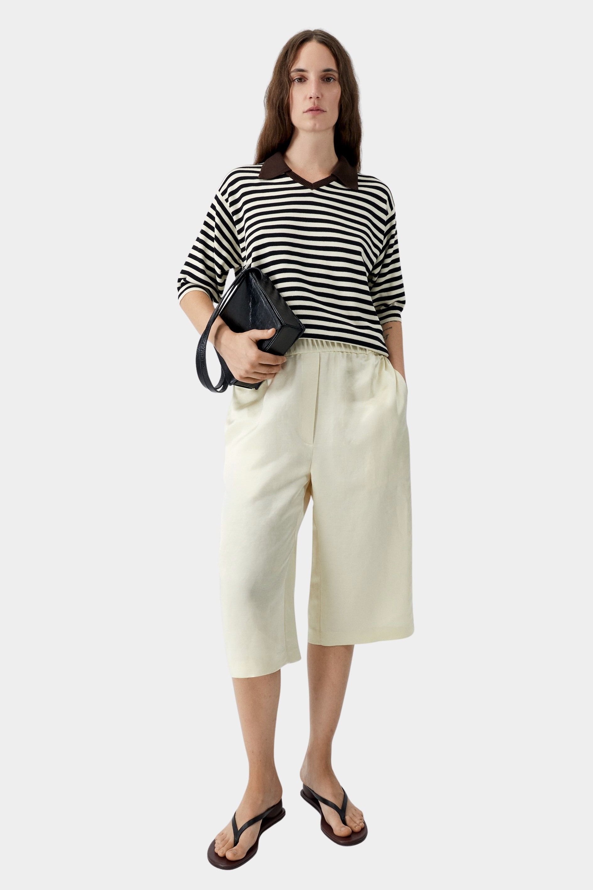 Cordera organic cotton V-neck polo in black and cream horizontal stripe with relaxed fit, short sleeves, and contrast brown collar, styled with cream culotte pants and black sandals