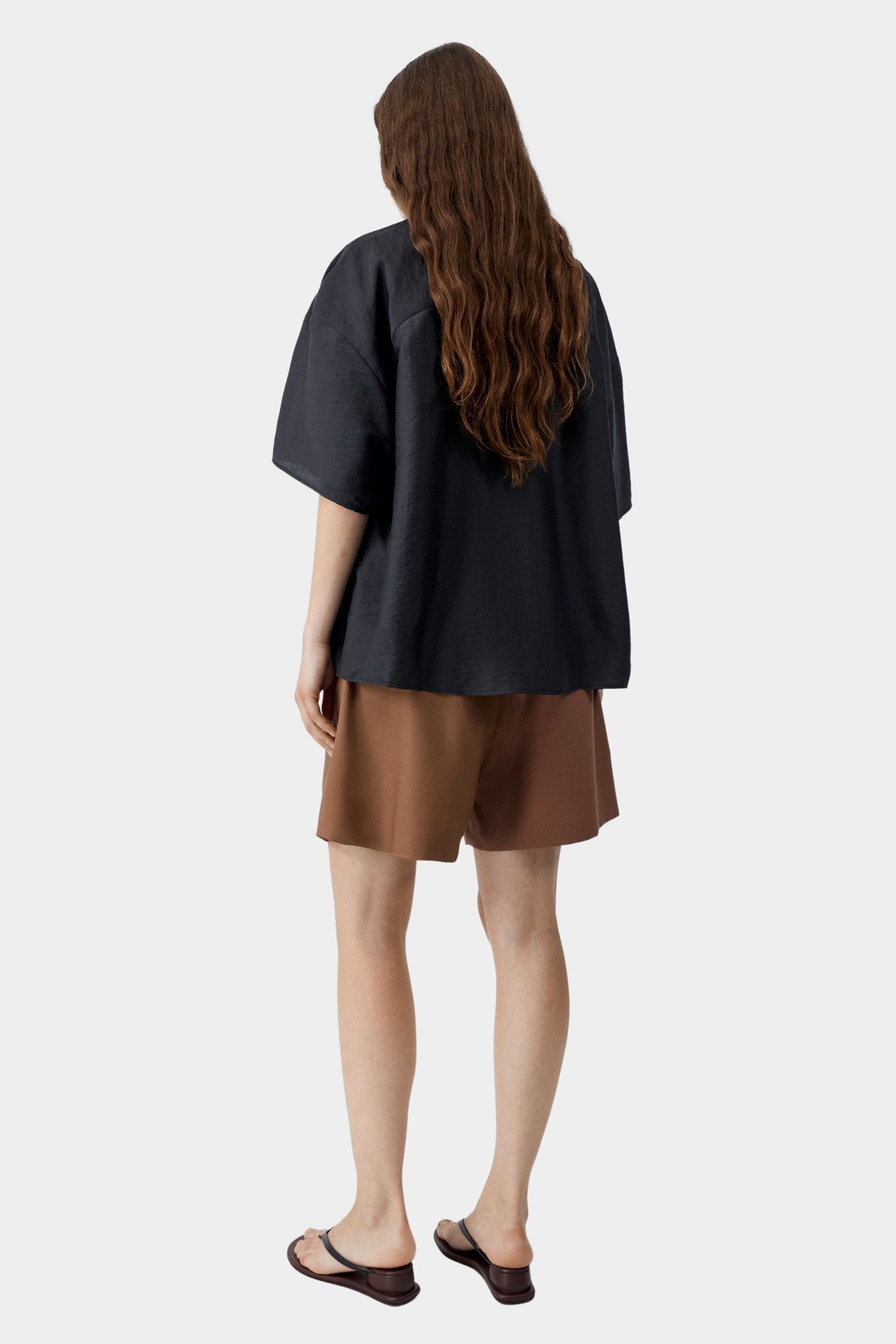 Black short sleeve shirt with relaxed oversized fit and clean back drape, worn with brown tailored shorts and sandals