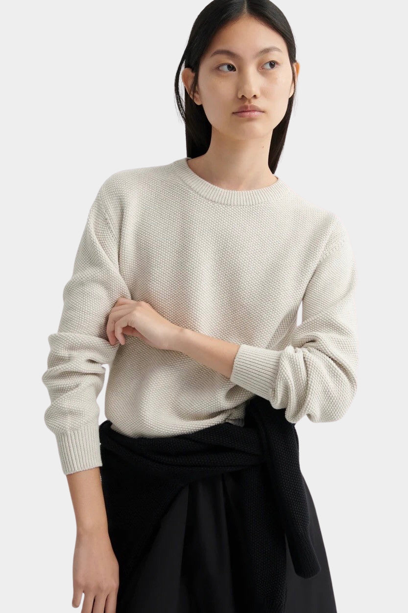 Model standing in a cream textured knit sweater with ribbed cuffs, styled with a black midi skirt.