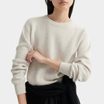 Model standing in a cream textured knit sweater with ribbed cuffs, styled with a black midi skirt.