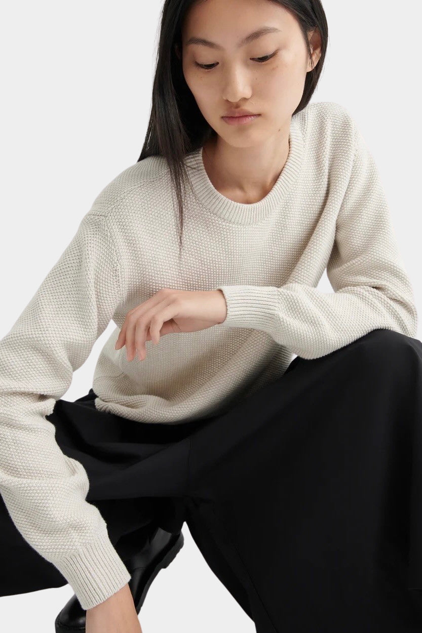Model seated wearing a textured cream crewneck sweater with black midi skirt and black leather shoes.