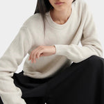 Model seated wearing a textured cream crewneck sweater with black midi skirt and black leather shoes.
