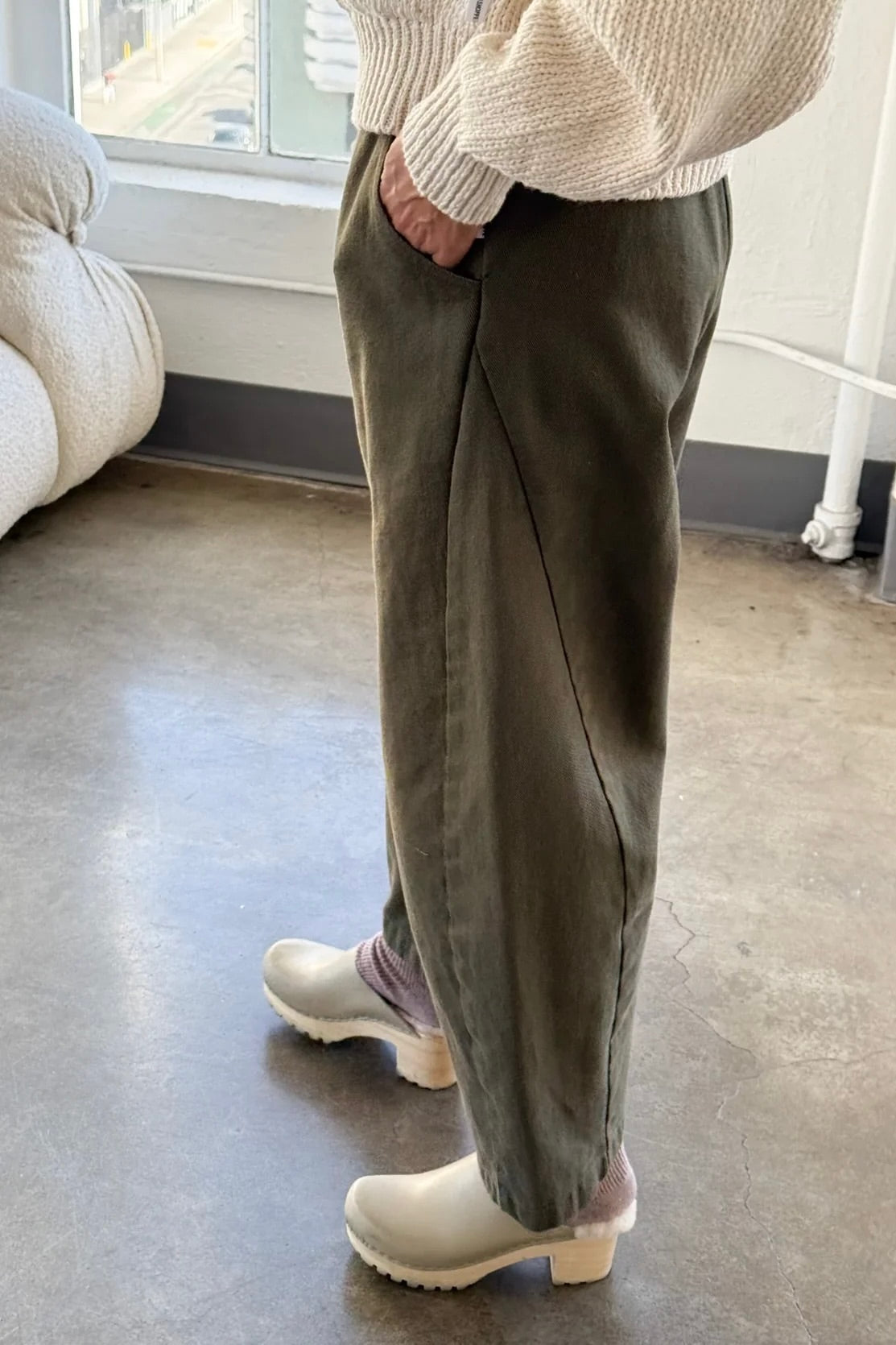 Side view of olive Arc Pants styled with a cream knit sweater and ivory clogs, highlighting the relaxed fit and curved leg silhouette.