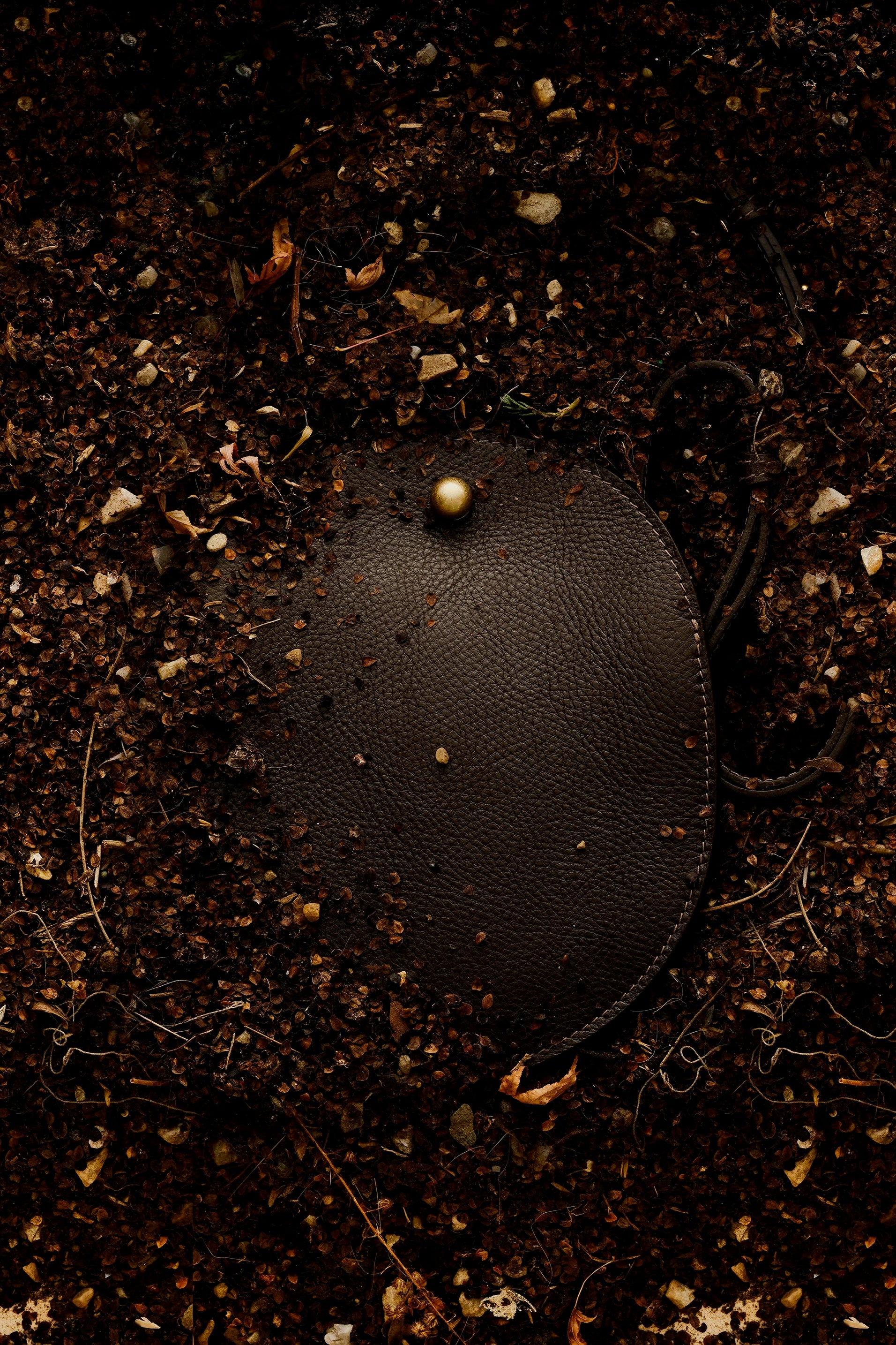 LINDQUIST Eggi bag in Chaga resting among soil and leaves, with pebbled leather and a spherical brass closure