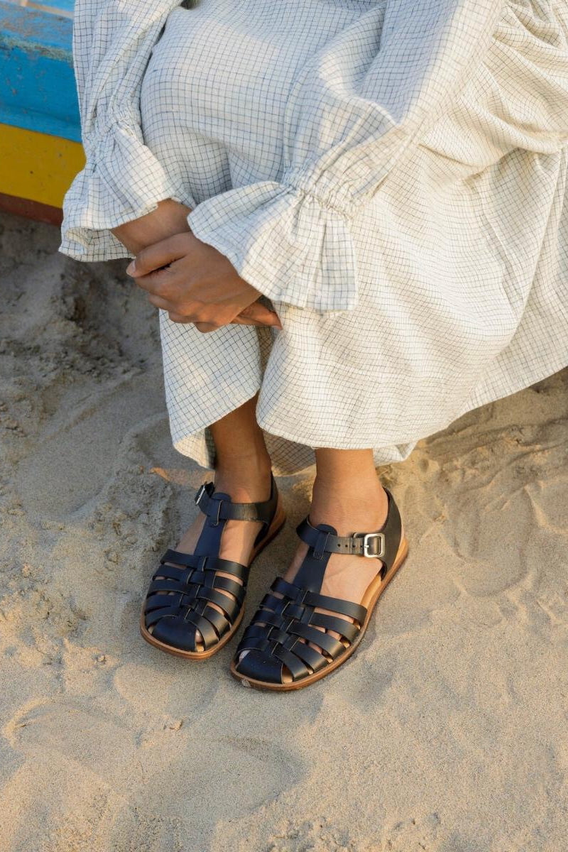 LOQ Rio sandal in black worn on sand with light dress showing flat leather fisherman sandal and ankle strap