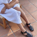 LOQ Rio sandal in black worn with white dress seated on tile floor showing fisherman sandal styling and buckle strap