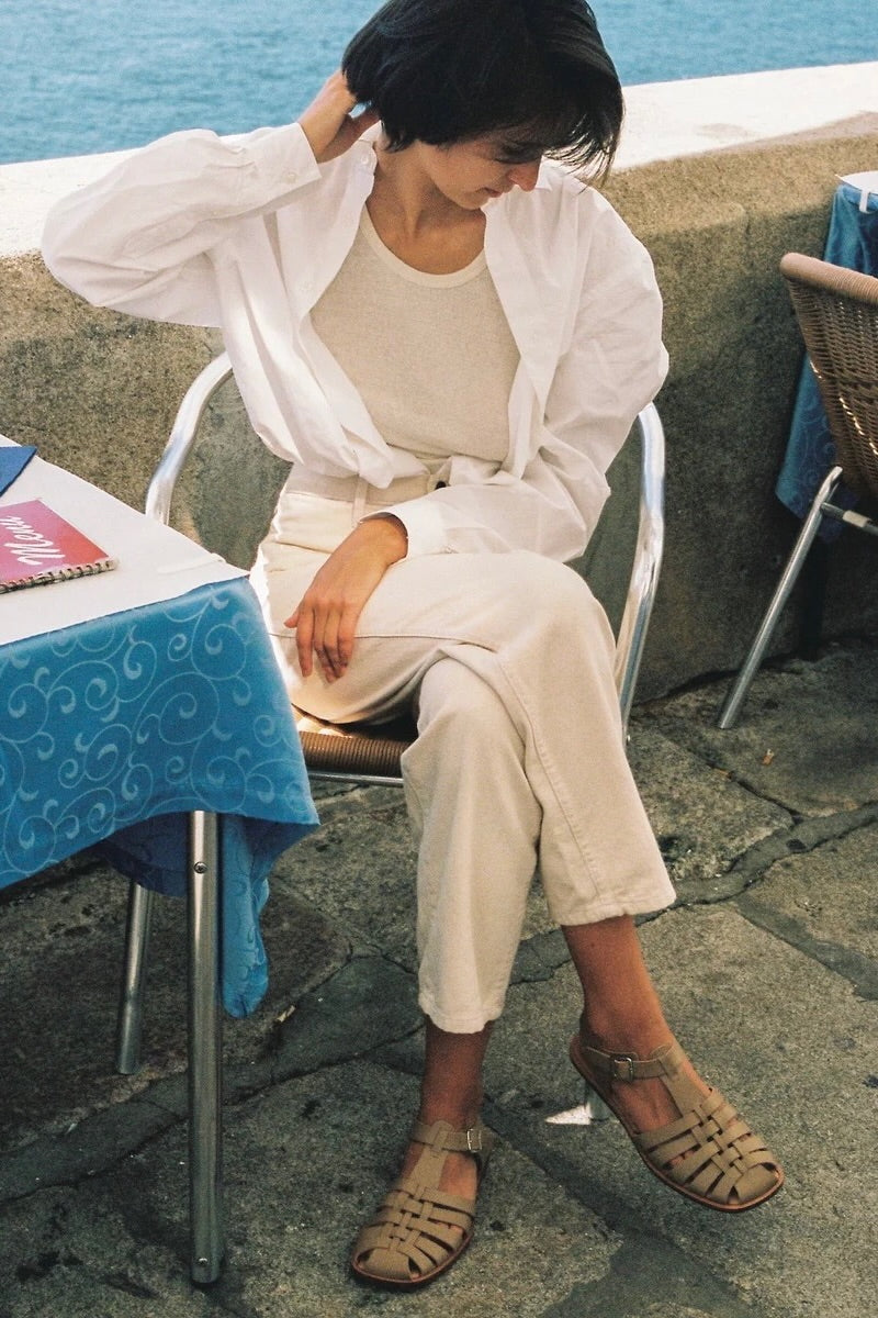 LOQ Rio sandal in taupe worn with cream outfit seated outdoor showing flat leather fisherman sandal and ankle strap