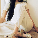 LOQ Rio sandal in taupe worn with cream pants seated indoor showing fisherman sandal styling and buckle strap