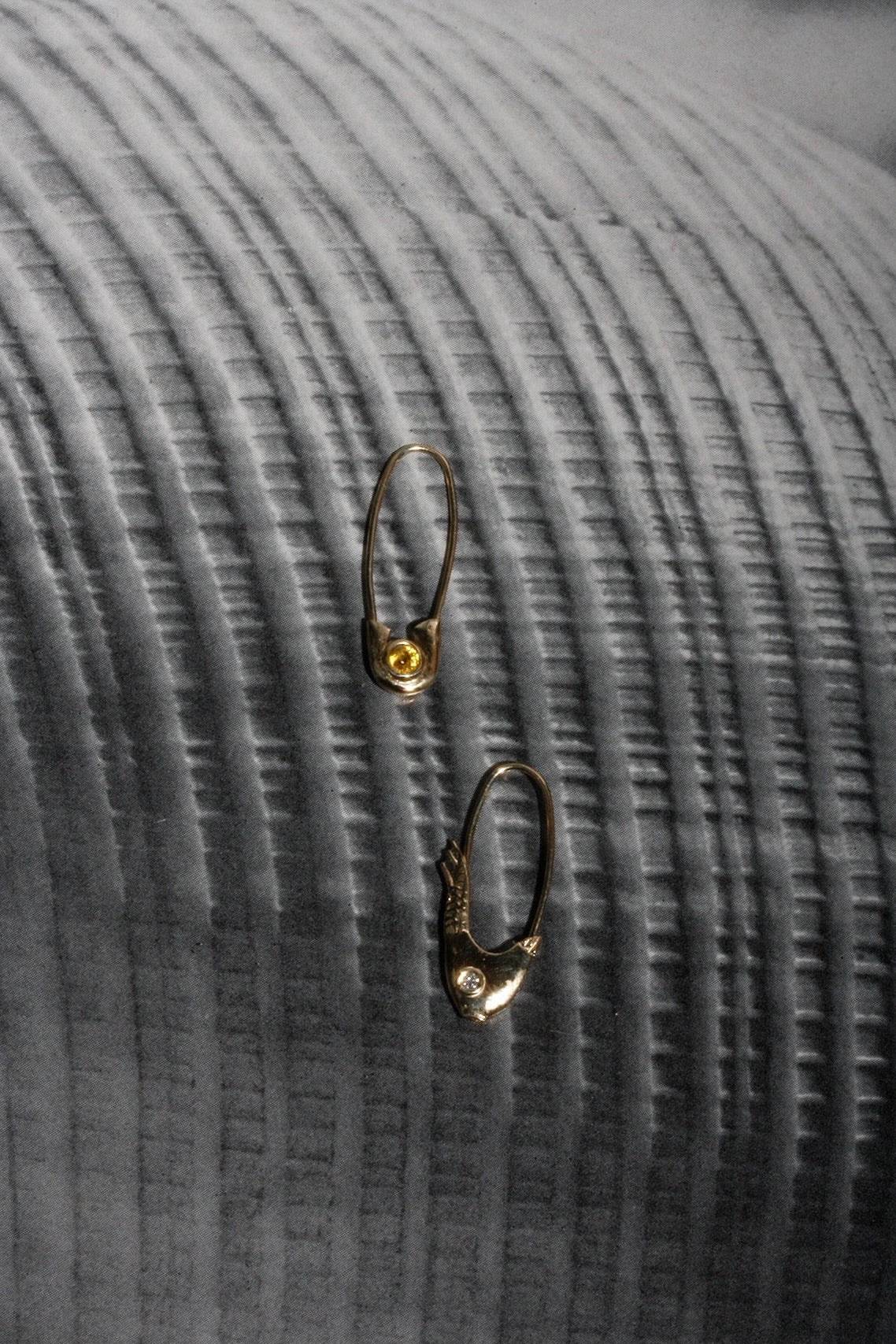 Two 14k yellow gold Pisces Safety Pin Earrings photographed on dark textured fabric
