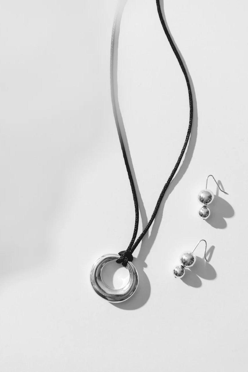 Maslo Jewelry sterling silver pendant necklace with circular ring charm on black cord styled with double orb silver drop earrings on light background