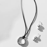 Maslo Jewelry sterling silver pendant necklace with circular ring charm on black cord styled with double orb silver drop earrings on light background