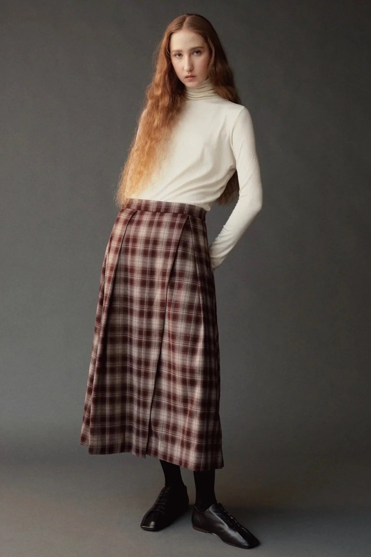 Model wearing the Mijeong Park brown plaid midi skirt with front pleats and an ivory turtleneck